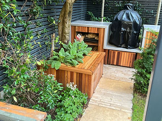 Cozy backyard corner with a wooden bench, lush green plants, and a covered barbecue grill.