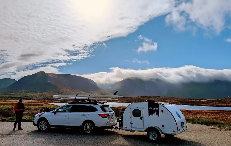 Igloo teardrop trailer towed by a station wagon loaded with paddleboards, in front of cloud-covered mountains