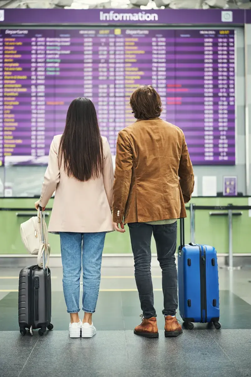 A couple standing with their luggage while looking at the flight departure information board at an airport.