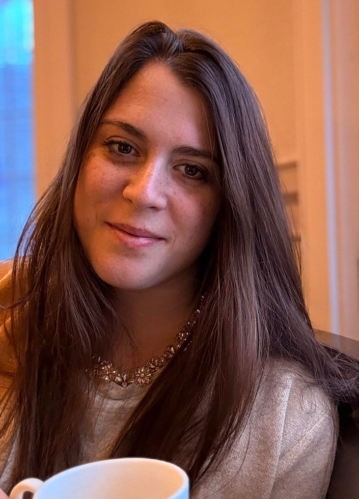 A young woman with long brown hair smiles subtly while sitting indoors near a window with a warm, ambient background in
