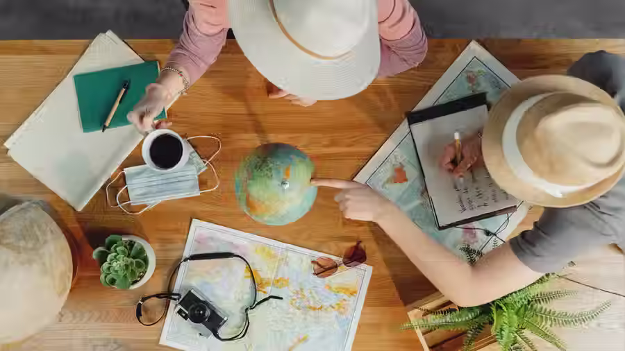 Overhead view of two people planning a vacation with a globe, maps, notebooks, and a camera on a wooden table.