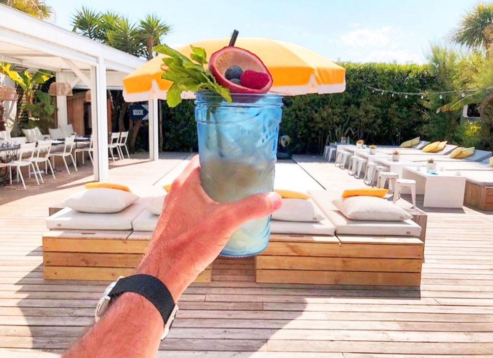 A hand holding an exotic and tropical cocktail under the sun with a sofa corner in the background