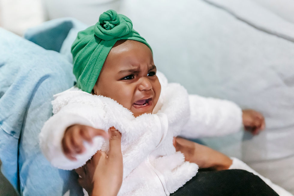 An upset baby in a green turban and fluffy white jacket being held and comforted by her mom on the couch