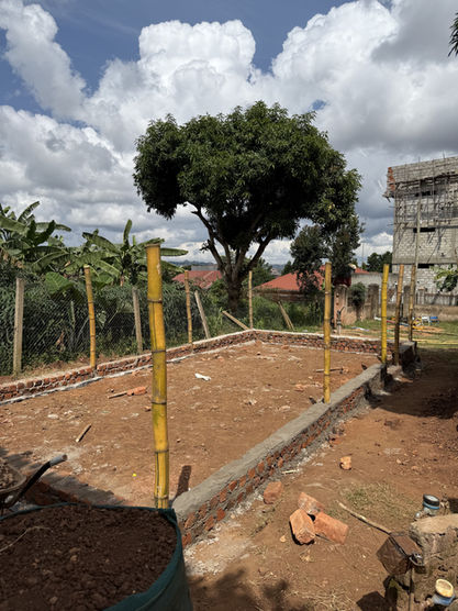 The concrete and brick foundation for a Walipini Impact Build is clearly visible, with sturdy bamboo poles erected as initial supports, illustrating the foundational steps of the Walipini Impact in Uganda