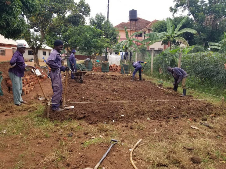 A team of Grace Place Volunteers in purple jumpsuits diligently prepare a plot of rich soil, using shovels and marking lines for a new Walipini structure, showcasing the foundational work for the Walipini Impact in Uganda