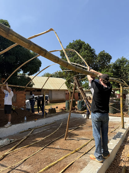 Grace Place Volunteers work together to construct the arched bamboo frame of a Walipini greenhouse in Uganda, securing long poles into a concrete base, demonstrating the hands-on building of the Walipini Impact in Uganda.	
