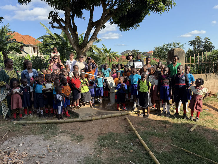 A large, joyful gathering of Ugandan Grace Place School children and several Grace Place members stand around a newly installed water pump under a large tree, celebrating improved access to water in their community, an essential step in supporting initiatives like the Walipini Impact in Uganda.	