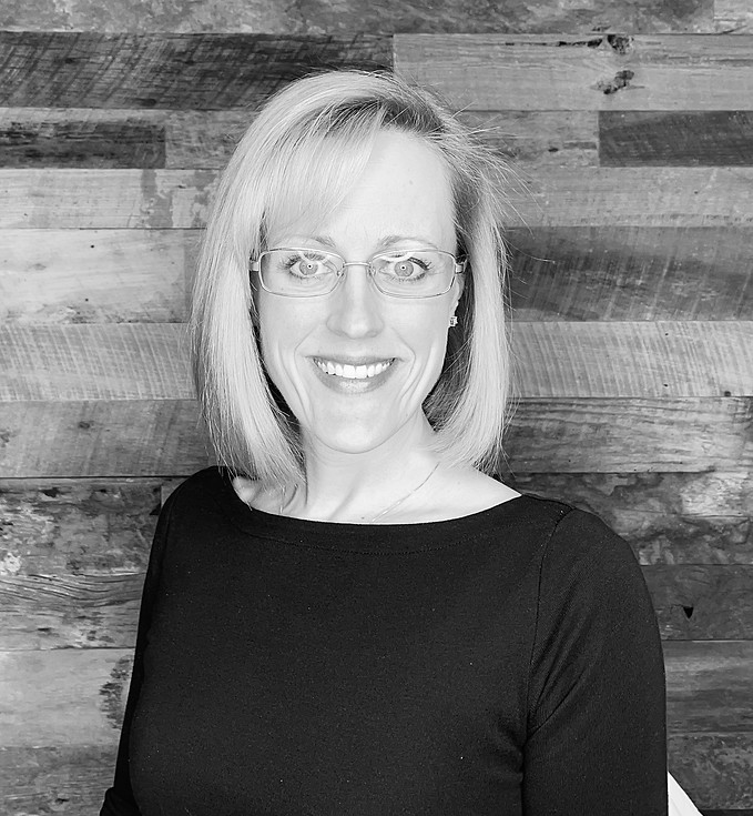 Black-and-white portrait of a smiling woman wearing glasses and a dark top, posed in front of a rustic wood plank wall.