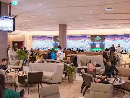 Spacious sports lounge with guests enjoying drinks and conversations, featuring large screens displaying a football game and comfortable seating arrangements.