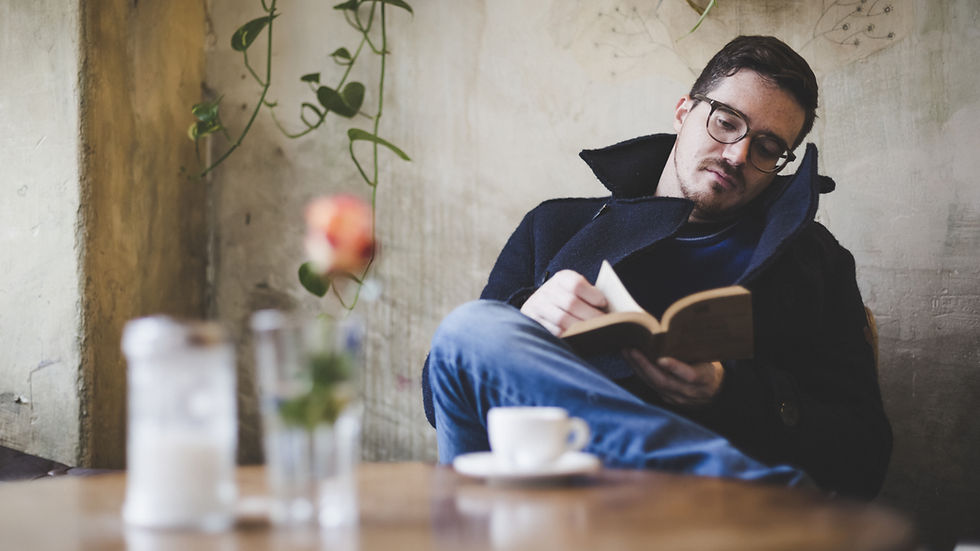 Man in glasses reading book, seated in cozy cafe with rustic walls and hanging plants; a blurred rose and coffee cup in foreground.