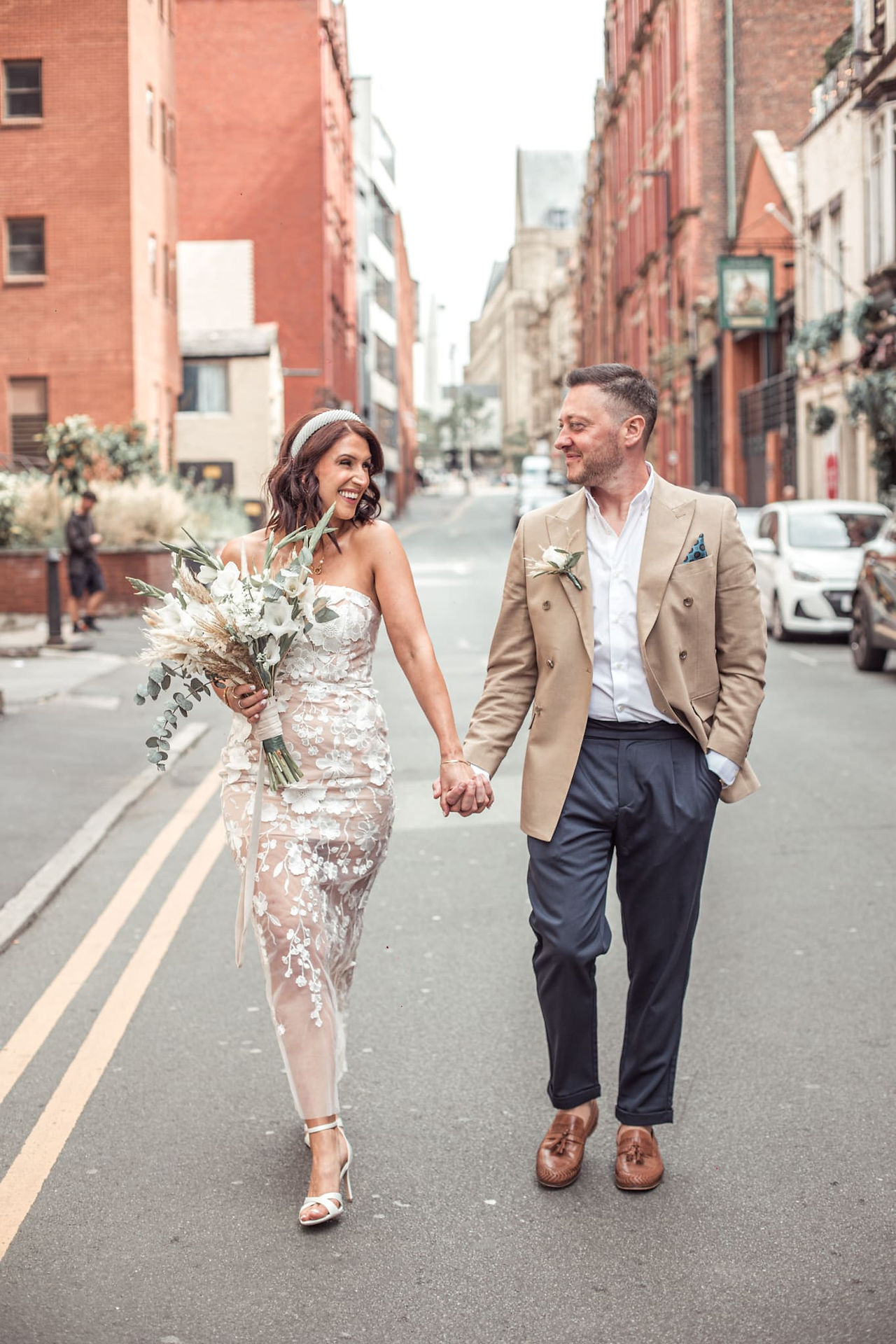 Bride and groom holding hands, standing in a Manchester street with red-brick buildings in the background – modern city wedding photography.