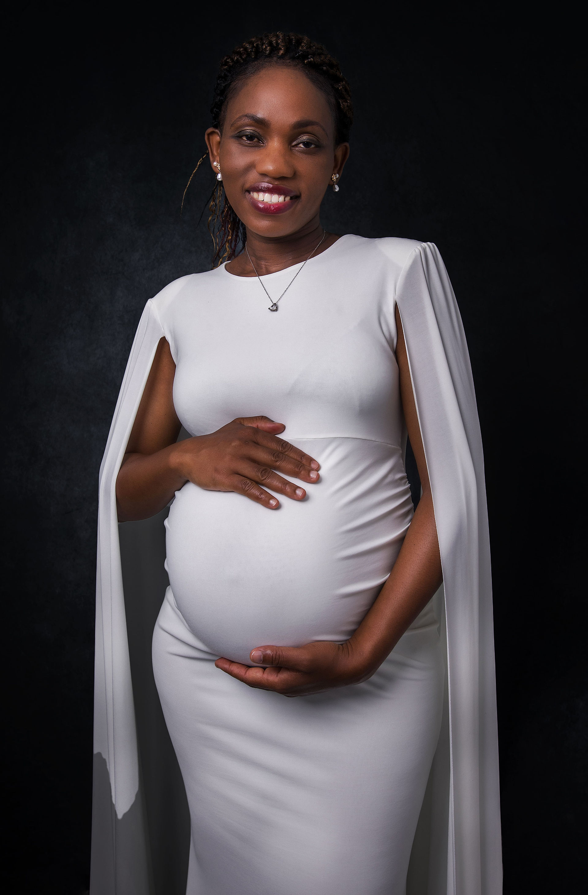 Pregnant woman gently holding her belly with both hands, smiling warmly while posing solo in her white maternity dress.