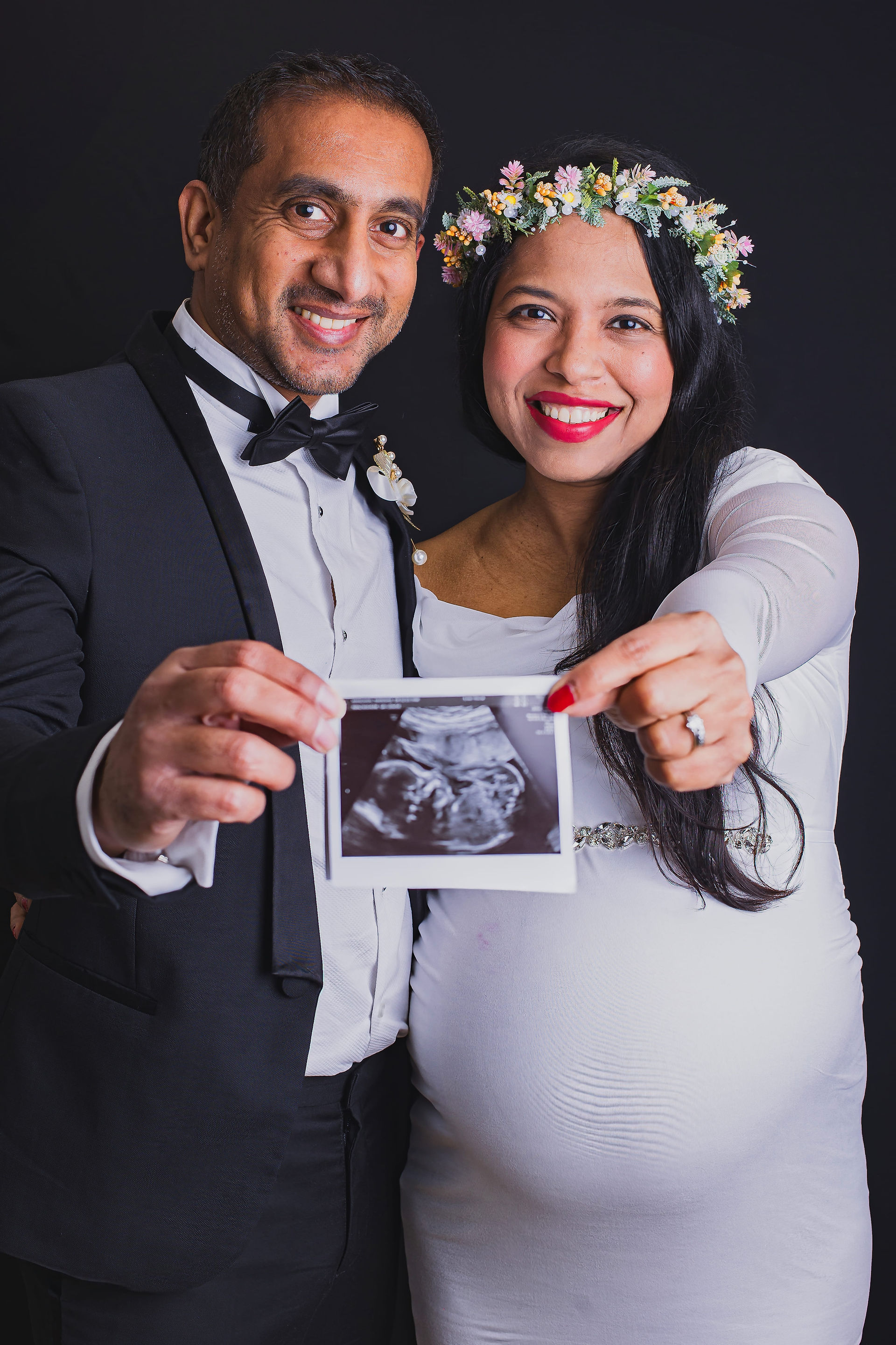 Expecting parents in formal wear smiling and holding an ultrasound photo toward the camera, highlighting their excitement and joy.
