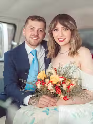 Smiling bride and groom sitting in the back of a vintage wedding campervan, with the bride holding a colorful bouquet.