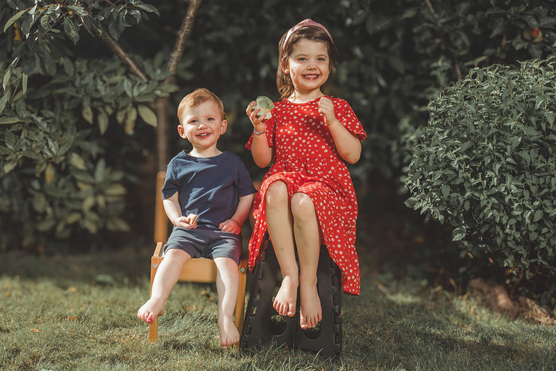 Young siblings sitting barefoot on stools in the garden, smiling and holding apples.