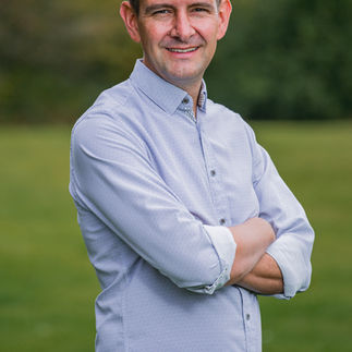 Friendly and approachable outdoor headshot of a man smiling with arms crossed, ideal for use on work websites and social media profiles.