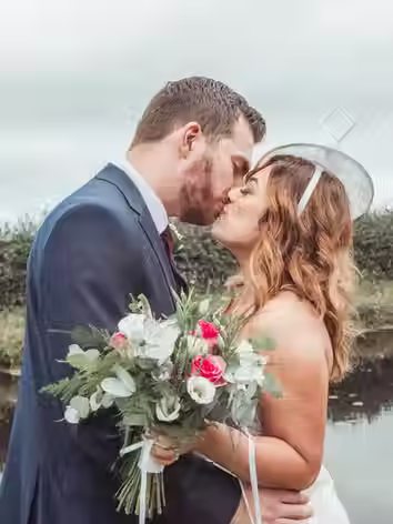 Close-up of a bride and groom sharing a kiss outdoors, with the bride holding a green and white floral bouquet