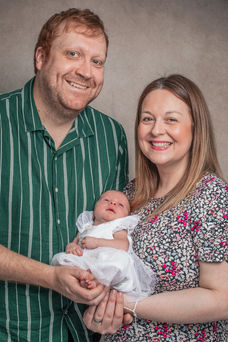 Smiling parents holding their newborn baby, posing in a family portrait with a neutral-toned backdrop, radiating love and happiness.