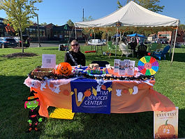 A woman sits at a table outdoors among green grass. Her table has the VSC banner and brochures about the agency.