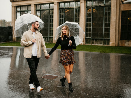 A smiling couple holding hands walks together in the rain, each holding a clear umbrella. The man wears a beige jacket and dark pants, while the woman is dressed in a floral skirt and black top. The setting is urban, featuring large windows and a reflective wet pavement.