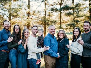 Extended family photo taken outdoors with golden morning light filtering through tall pine trees. Three generations stand together smiling, dressed in coordinated jewel and neutral tones. Photographed by Laura Patrick Photography.