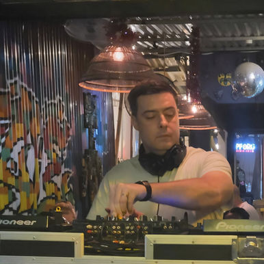British Drum and Bass DJ Loxley performing at an event in a venue on Pioneer CDJs, looking at the left CDJ in a colour picture taken from his front.