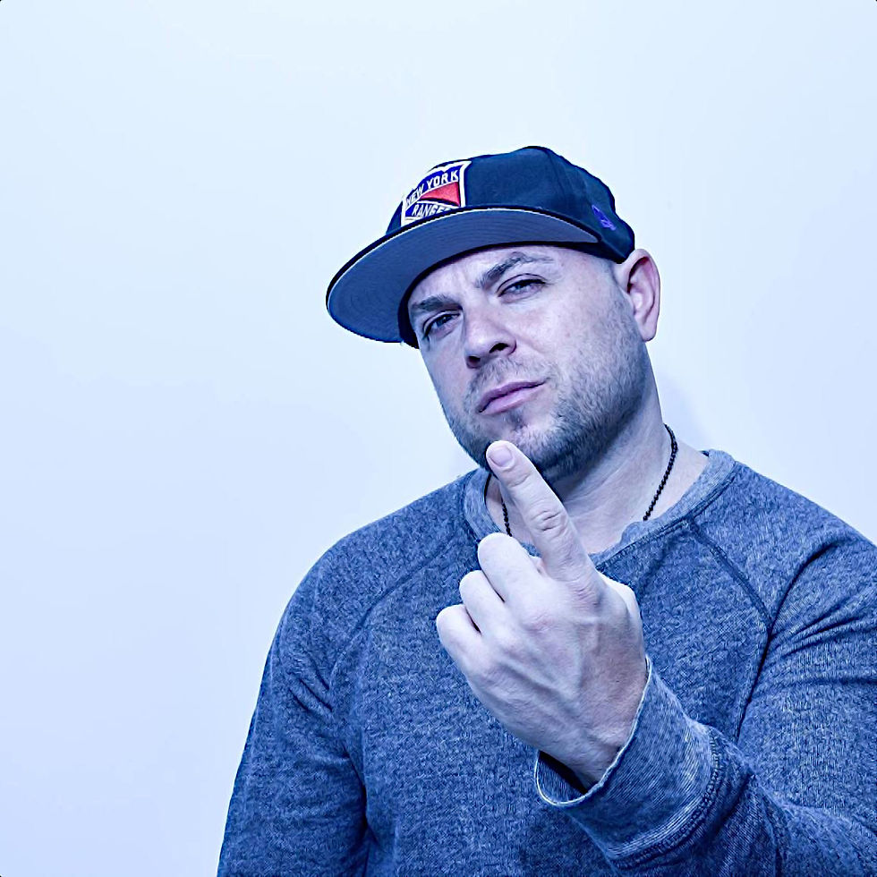 American Lyricist and rapper El Gant wearing a baseball cap with his right hand raised and his index finger pointing upwards, smiling wrly at the camera in an upper body shot, colour image.