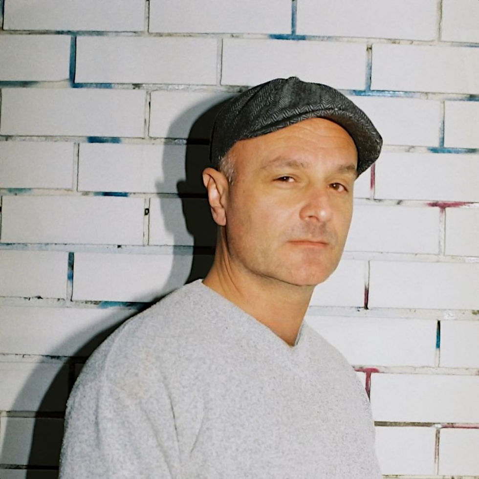 Australian DJ Paul Cazzar standing wearing a flat cap in front of a painted brick wall and looking slightly over his right shoulder at the camera, colour image.