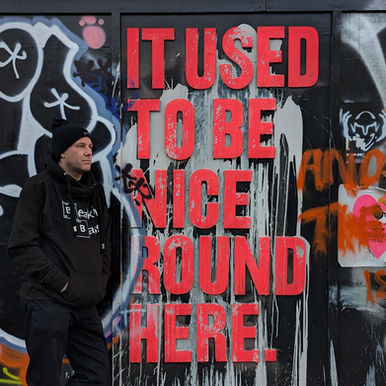 Blaze DJ standing in London beside a graffiti-covered wall featuring large red text reading “It Used To Be Nice Round Here,” wearing a black hoodie and beanie.