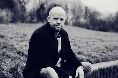Black and white portrait of German producer Roland Leesker, seated outdoors, wearing a dark jacket.