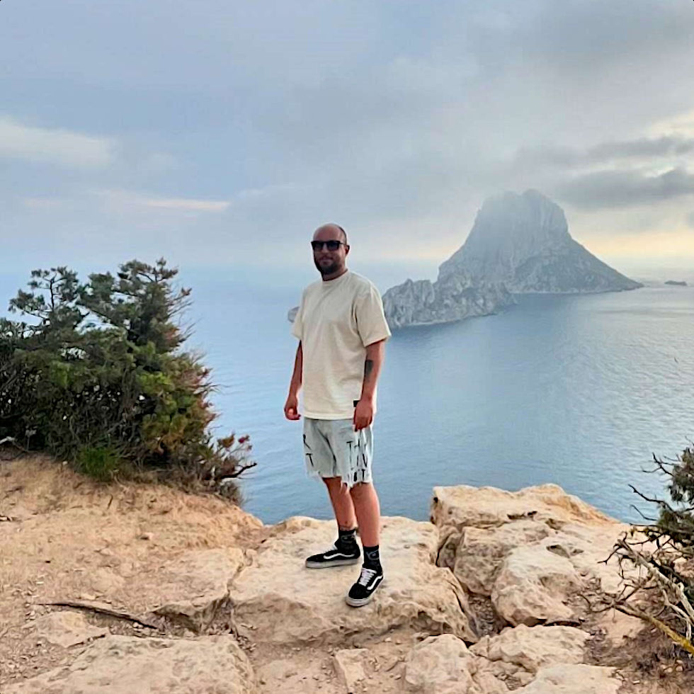 Spanish DJ and producer RaMuco standing on a rock next to the sea in Ibiza, Spain, colour image.