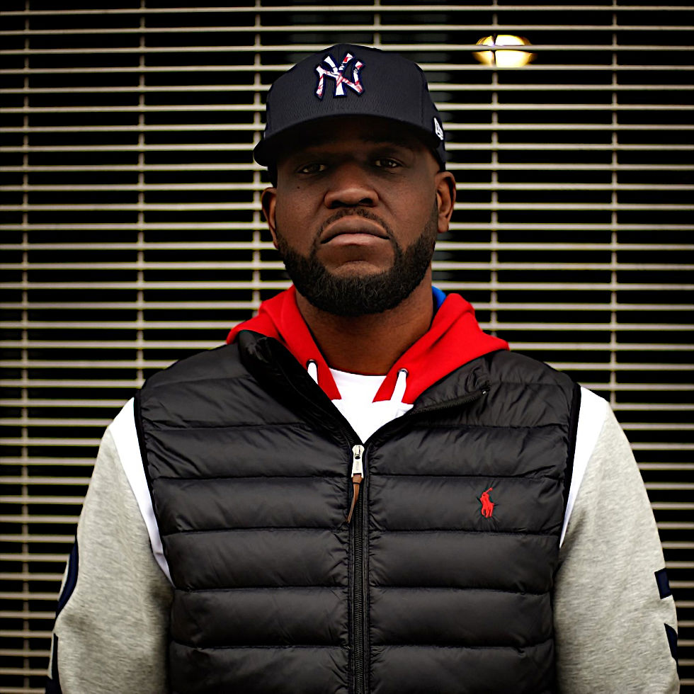 Canadian Rapper and producer Connaisseur Ticaso standing in upper body shot looking at camera wearing a baseball cap and sleeveless padded jacket over a hoodie, colour image.
