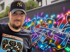 French DJ and Videographer DJ Phoenixx standing next to Graffiti wall with his right hand raised in a two fingered gun salute wearing a Baseball Cap and T shirt, colour image