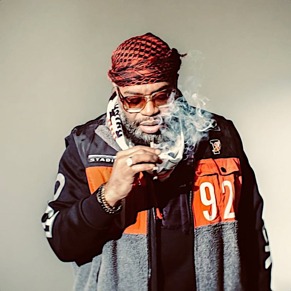 American Rapper and producer D.V Alias Khryst wearing a jacket, sunglasses, and a shemagh and smoking a joint whilst looking down at the ground, colour image.