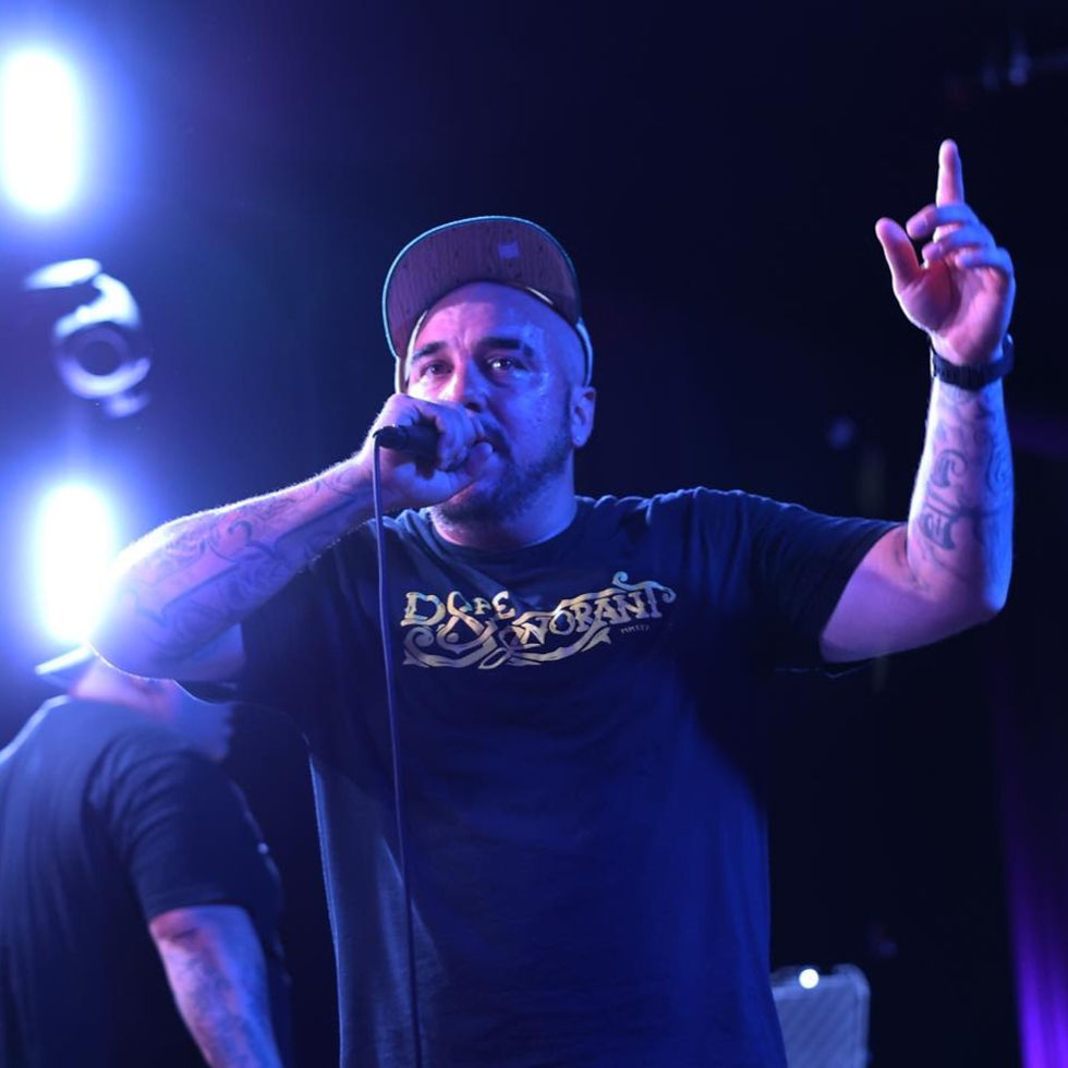 M-Dot performing live on stage under blue lighting, holding a microphone in one hand and raising the other, wearing a black T-shirt and cap.