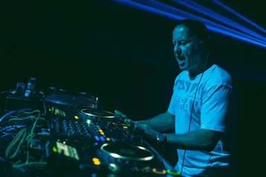 DJ Graham Gold performs behind a club DJ setup under blue lighting, captured mid-set with headphones on and stage lasers in the background.