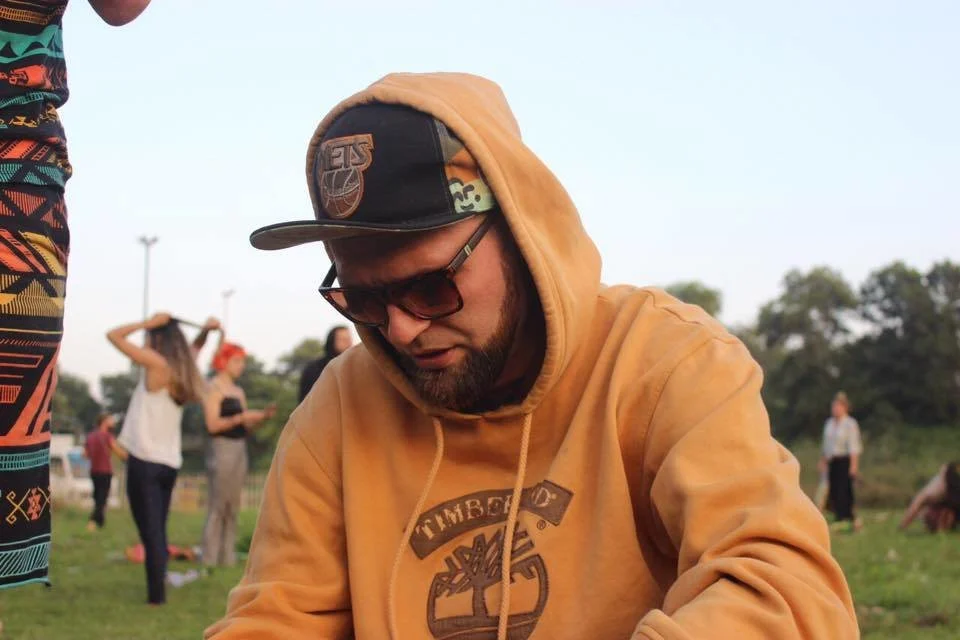 Russian DJ and MC 8U8A sitting on grass at festival wering sunglasses and a baseball cap with hoodie on, colour image.