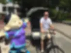 Italian DJ and producer Marco Mei sitting on a cycle rickshaw, pretending to pedal with a large cartoon statue of a woman next to him, colour image.