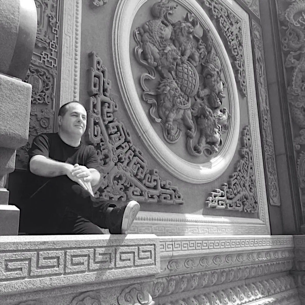 Italian DJ and producer Marco Mei sitting on a pliunth of a wall with chinese carvings in the stone in the wall, black and white image.