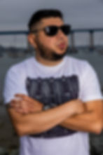 American DJ and radio presenter DJ Nocturne is portrayed in a lively scene by the harbor, gazing to his left with arms crossed. He sports a white t-shirt emblazoned with a Pioneer DJM800 mixer design and a pair of sunglasses, set against the bustling backdrop of the waterfront.