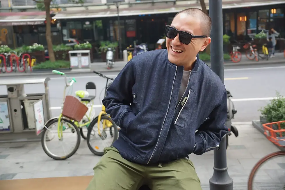 Taiwanese DJ and producer Murphy Kin a.k.a DJ lucy sitting on a bench and laughing wearing sunglasses in front of a street, colour image.