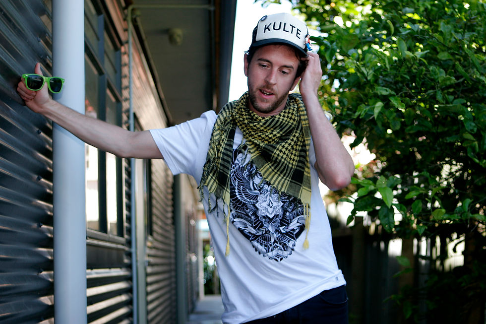 Jstar leaning on a building with sunglasses in his hand, colour image.