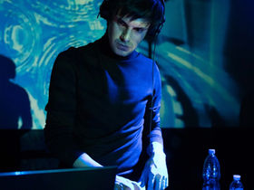 DJ Lastveek performing a live set in a club setting with visual projections and blue lighting.