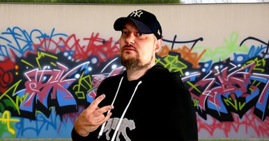 DJ Pheonixx standing in front of graffiti wall with two fingered pistol salute, colour image.