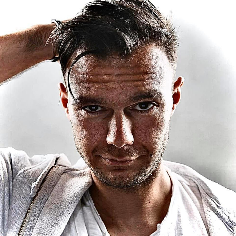 Italian DJ and Producer Andrea Mirgone standing with his hands in his pockets and smiling at camera in with his right hand behind his head, colour image.