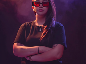 Latin American DJ Cinthia G standing with arms crossed, wearing red sunglasses and a black shirt, lit by purple and red studio lighting against a dark background.