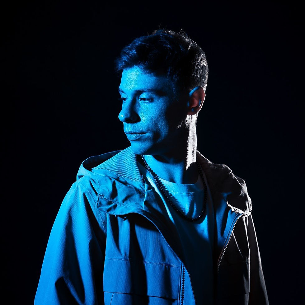 Argentinian DJ and producer Ivan Lozano is standing in a photo studio and looking to his right with blue lighting on him with a black background in a colour image.
