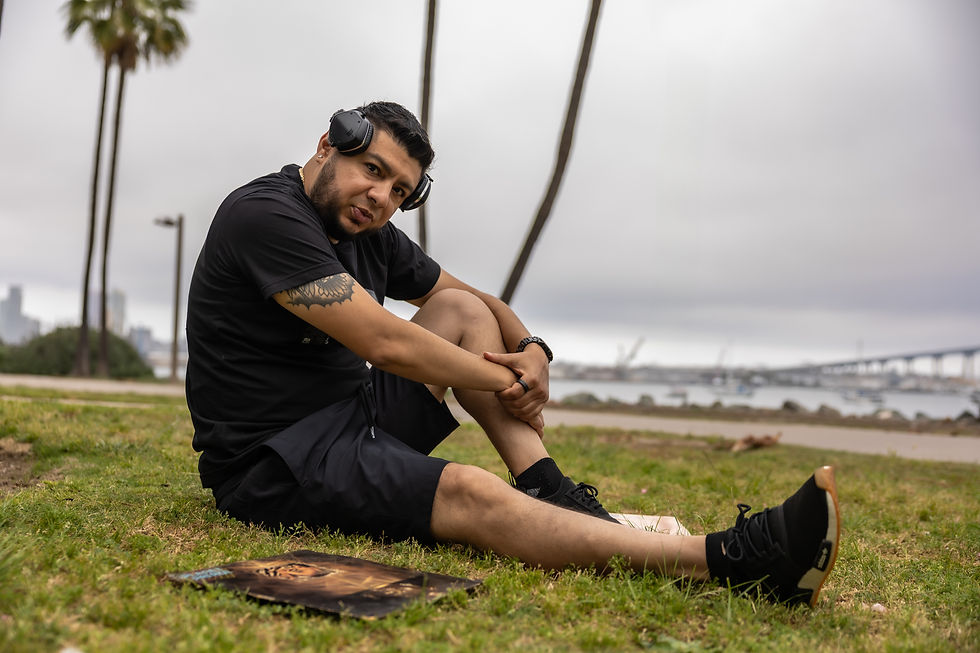 DJ Nocturne sitting on the grass by the waterfront in Los Angeles, wearing headphones, with the harbour bridge and city skyline visible in the background in a colour image.