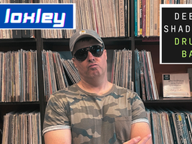 British Drum and Bass DJ Loxley is pictured standing in front of his record collection, wearing a camouflage T-shirt, aviator sunglasses, and a baseball cap, in a front-facing shot. The vibrant image displays his name in white on a blue background, along with the phrase "Deeper Shades Drum and Bass" set against a black background.