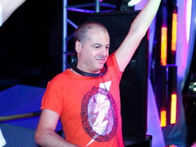 Italian DJ and producer Marco Mei performing at Geomatria with his left arm extended in a salute to the crowd and smiling profusely in a colourful image.